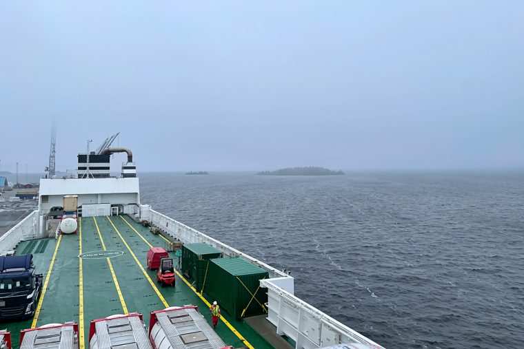 Bilden visar en gråmulen horisont och regntunga moln. I horisonten syns två öar. I förgrunden syns lastningen på ett fartyg.