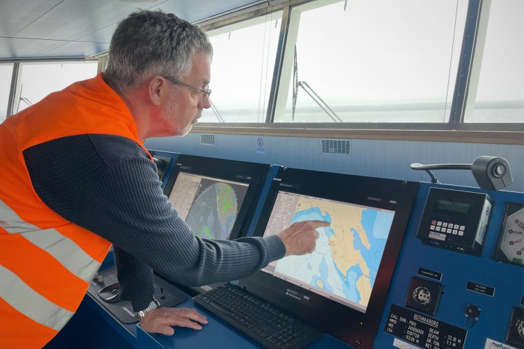 Bilden visar en man som står ombord på lastbryggan i ett fartyg. Mannen pekar mot en datorskärm som visar kartbilder.